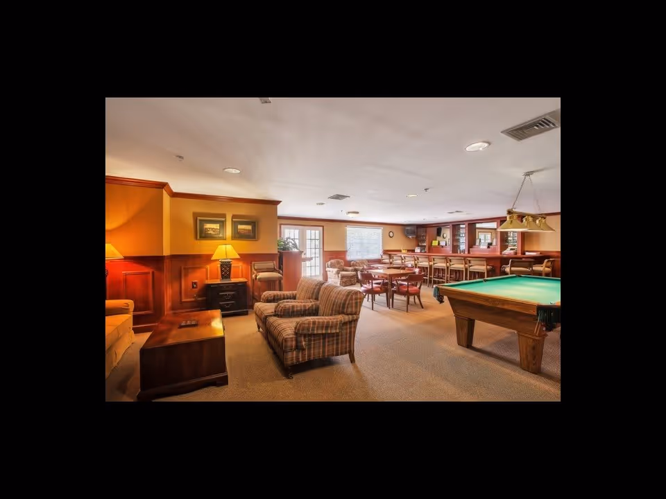 A spacious common room with comfortable plaid armchairs, a wooden coffee table, a pool table with green felt, and multiple tables and chairs for dining or socializing. The room has warm lighting, wood paneling on the walls, framed pictures, and large windows letting in natural light.