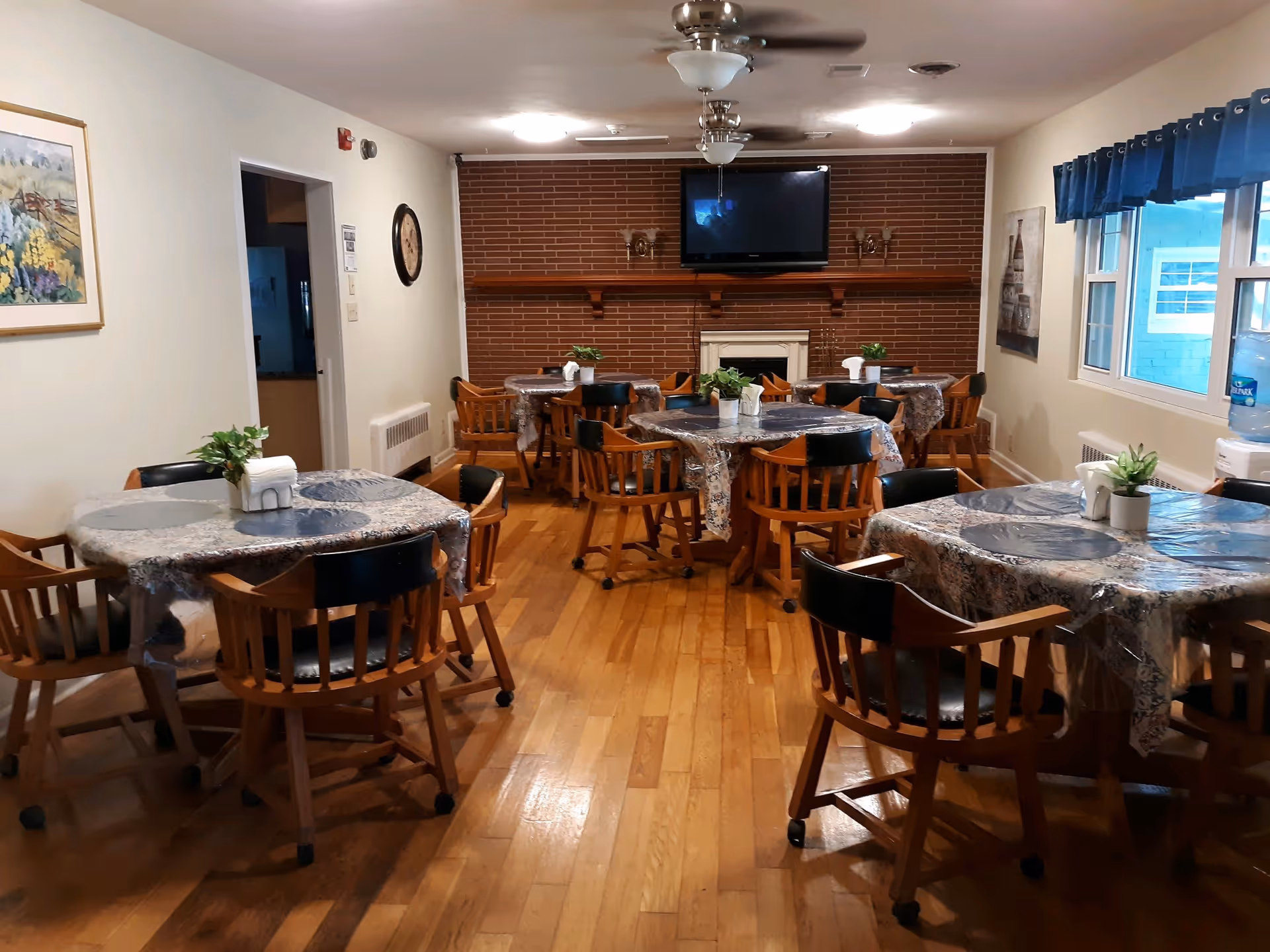A dining room with several round tables covered with patterned tablecloths and small potted plants as centerpieces. Wooden chairs with black seats surround the tables. The room has hardwood floors, a brick wall with a mounted flat-screen TV above a white fireplace, ceiling fans with lights, and windows with blue valances on the right side. There is a water dispenser near the windows and framed artwork on the walls.