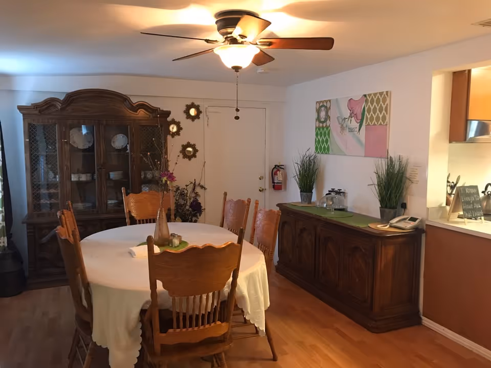 A dining room with a round table covered in a white tablecloth surrounded by wooden chairs, a wooden china cabinet and sideboard, ceiling fan, and wall decor.