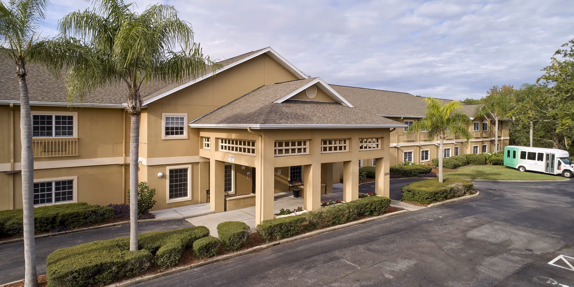 Front entrance of a two-story tan senior living building with palm trees and a green shuttle bus parked nearby.