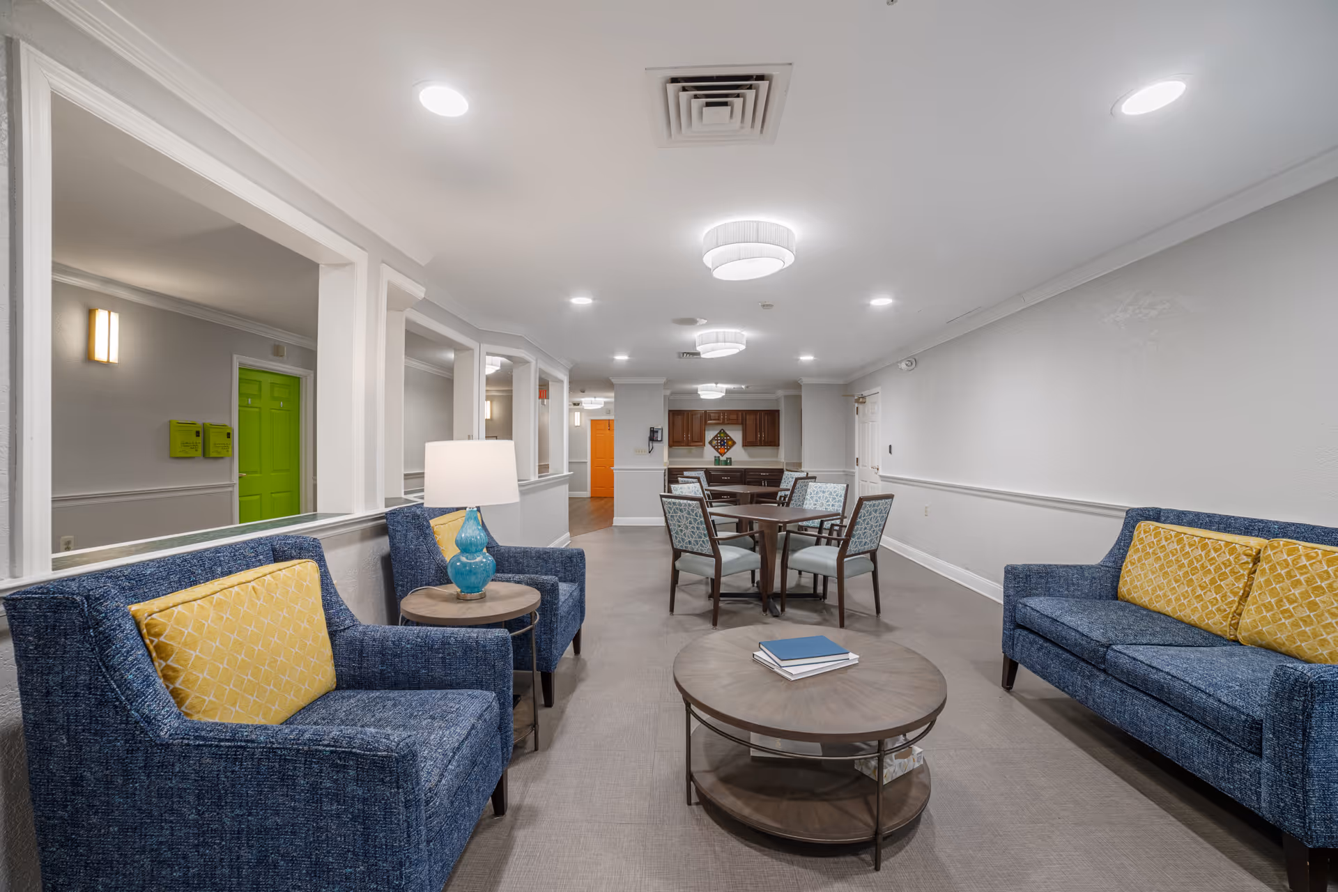 A well-lit interior common area with blue upholstered armchairs and a sofa with yellow cushions, a round wooden coffee table with books, a side table with a blue lamp, and a dining table with four chairs in the background. The walls are light gray with white trim, and there are green and orange doors visible in the hallway.