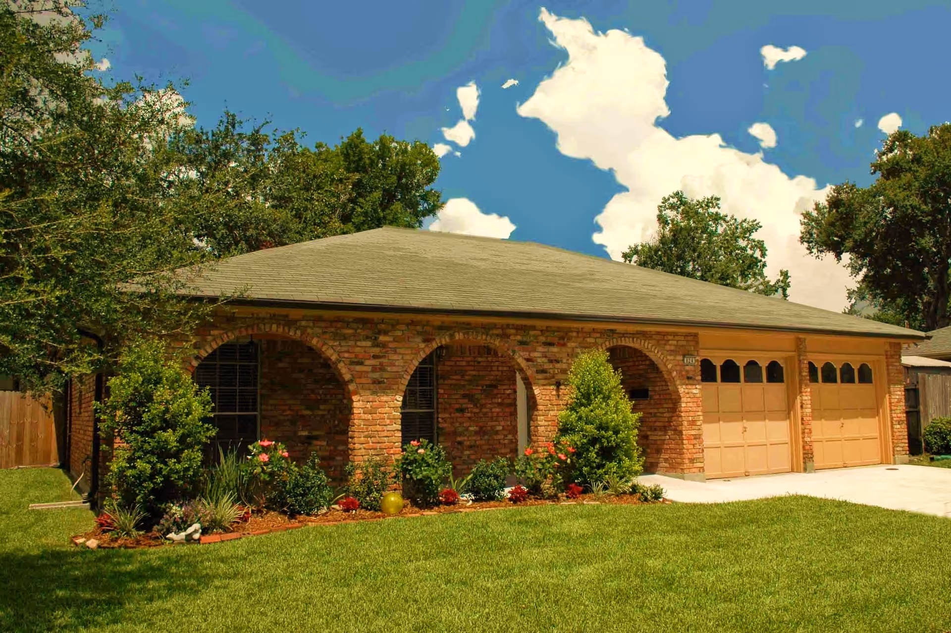 Single-story brick house with a green lawn, three arched front porch openings, and a double garage door under a clear blue sky with some clouds. The house is surrounded by trees and shrubs.