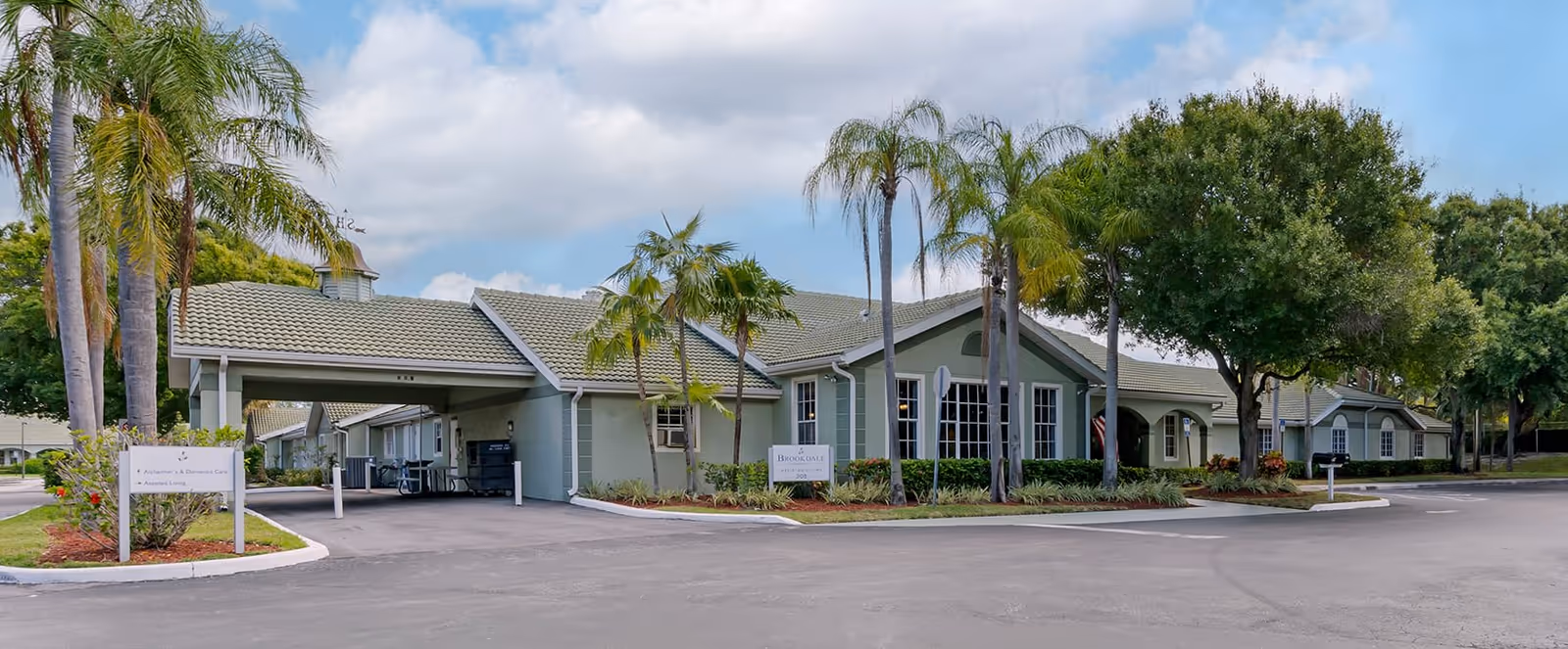 Exterior view of Brookdale Tequesta senior living facility showing a single-story building with a covered entrance, surrounded by palm trees and other greenery under a partly cloudy sky.