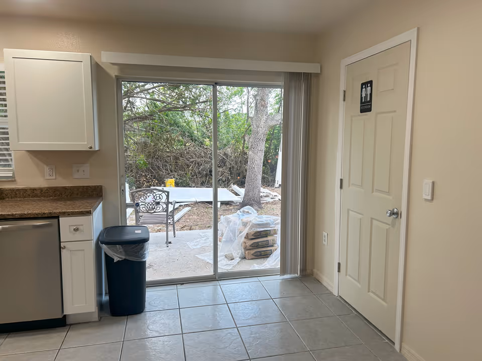 Small kitchen area with cabinets, dishwasher, trash can, and a sliding glass door opening to an outdoor patio, with a door marked as a restroom on the right.