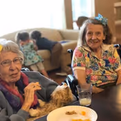 Two elderly women sitting at a table indoors, one holding a small dog and the other smiling, with a plate of snacks and a glass of milk on the table. In the background, two children are sitting on a couch near a window.