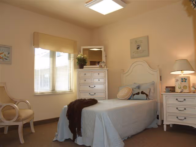 A cozy bedroom with a single bed covered in a light blue quilt and decorative pillows, a white wooden headboard, a matching white dresser with a mirror and a plant on top, a white nightstand with a lamp and decorative items, a beige armchair, and a window with blinds and a beige valance letting in natural light.