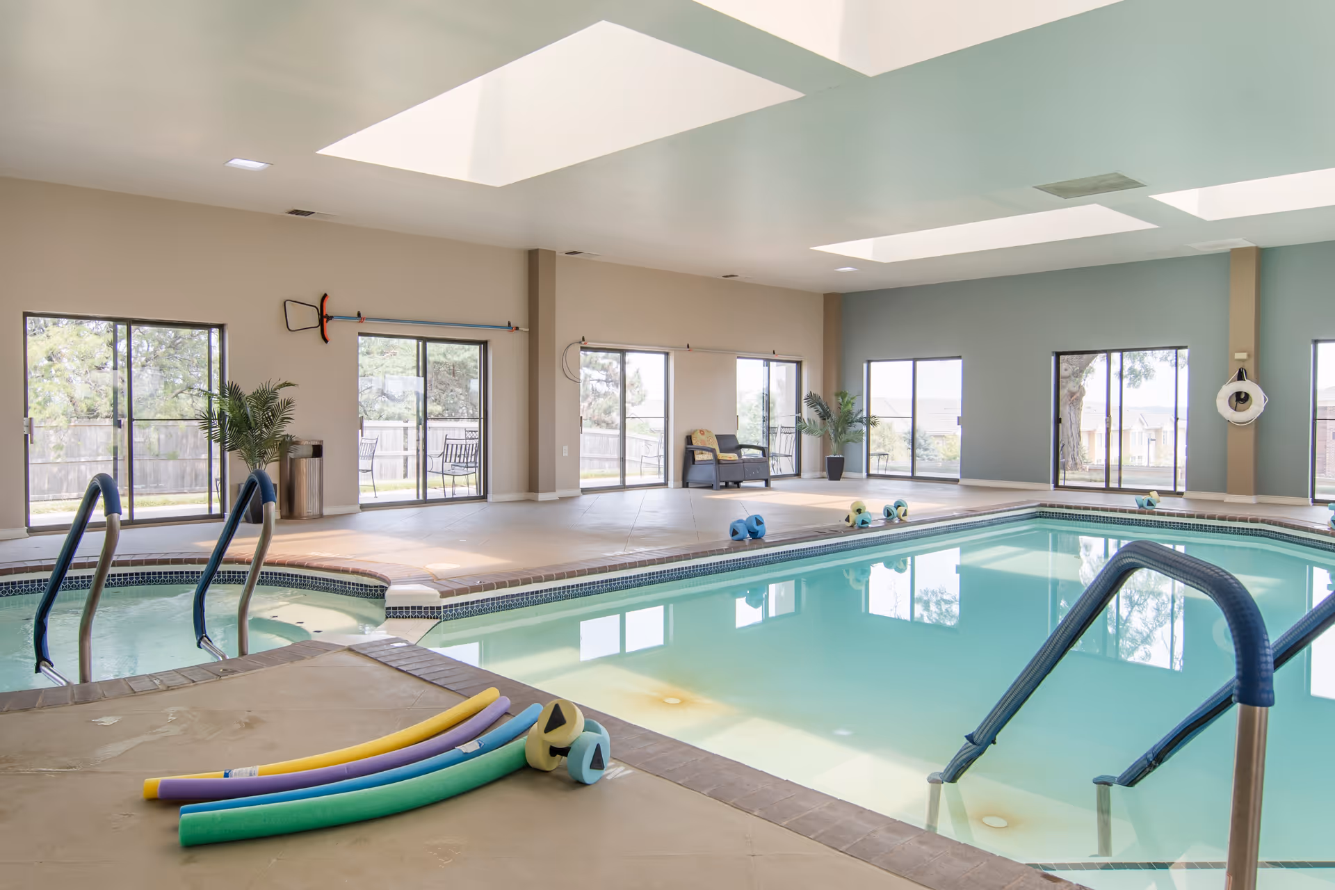 Indoor swimming pool area with large windows letting in natural light. The pool has handrails and pool noodles placed on the edge. There are chairs and plants around the pool area, and the ceiling has skylights.