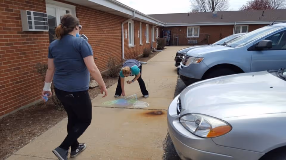 Two women drawing colorful chalk art on a sidewalk outside a single-story brick healthcare building beside parked cars.