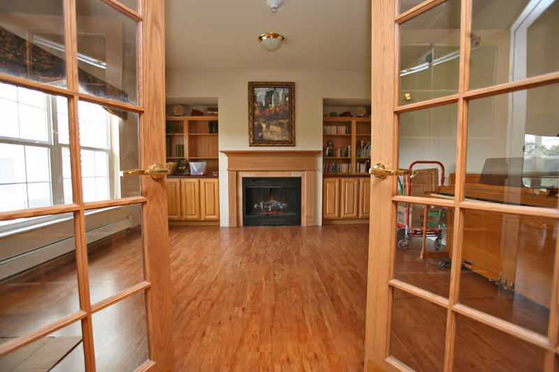 View through open wooden French doors into a room with wooden flooring, a fireplace centered on the far wall, built-in wooden bookshelves on either side of the fireplace, and a painting hanging above the fireplace. Large windows on the left side allow natural light to fill the room.
