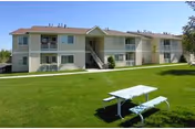 Two-story apartment building with beige siding and balconies, surrounded by a well-maintained green lawn with a white picnic table and benches in the foreground under a clear blue sky.