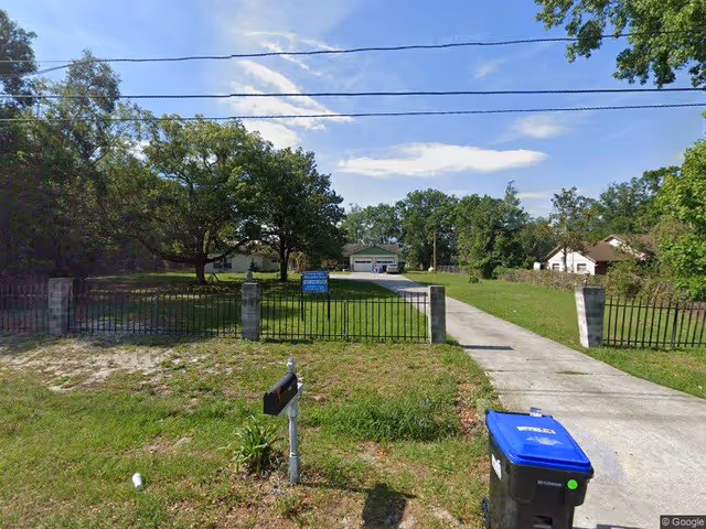 View of a residential property with a driveway leading to a garage, surrounded by trees and grass. There is a black metal fence with stone pillars at the entrance, a mailbox near the road, and a blue recycling bin on the right side of the driveway. The sky is partly cloudy.