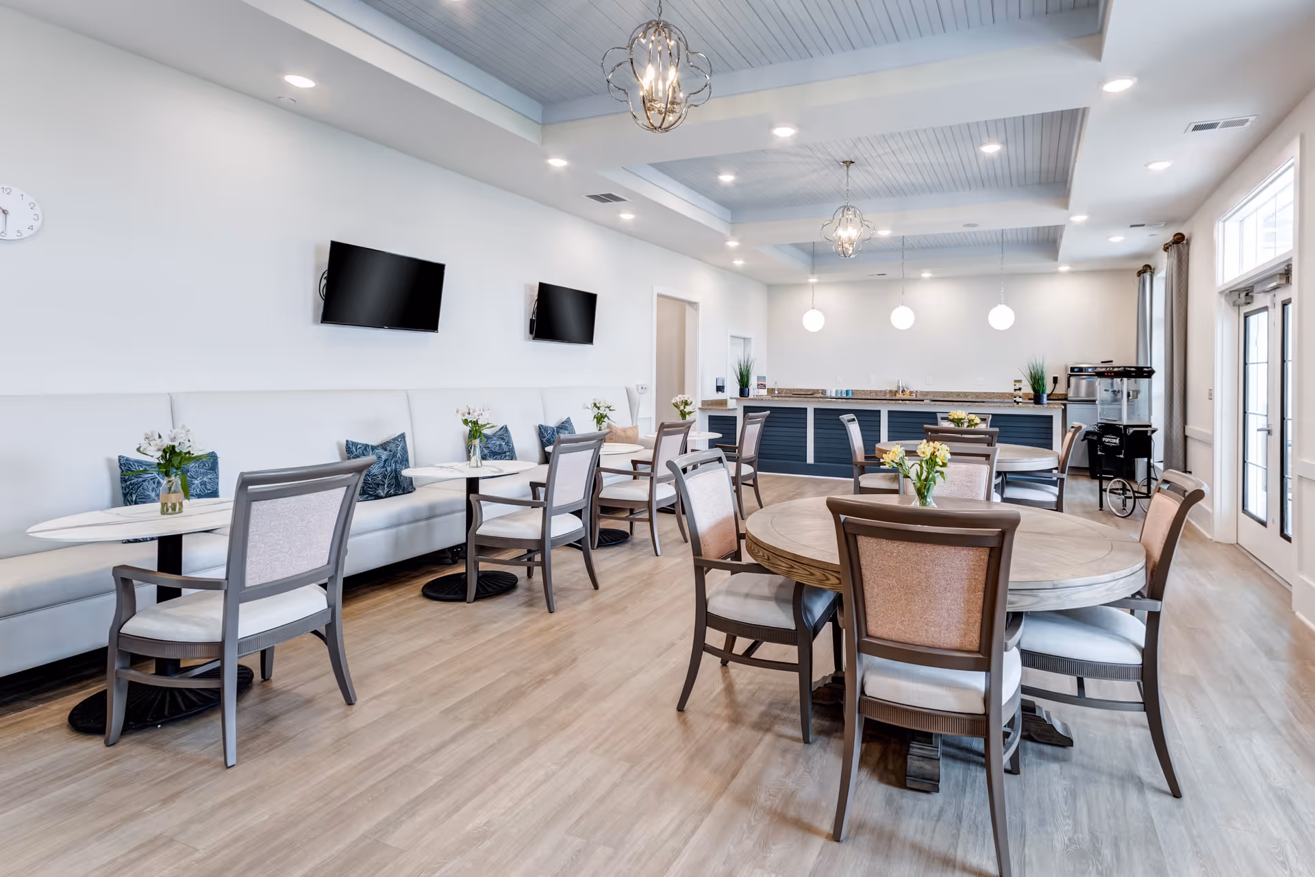 A bright and modern dining room with multiple round and rectangular tables surrounded by chairs. The room features light wood flooring, white walls, and a ceiling with recessed lighting and decorative hanging light fixtures. There are two flat-screen TVs mounted on one wall above a long cushioned bench with decorative pillows and small flower vases on the tables. At the back, there is a counter area with a granite countertop, plants, and a popcorn machine near the door.