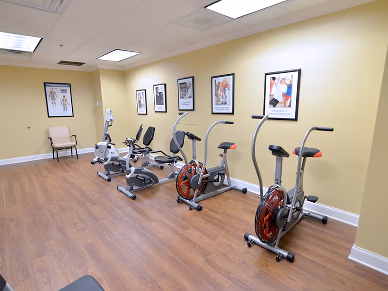 A small exercise room with wooden flooring and beige walls featuring four stationary exercise bikes lined up against the wall. There are framed motivational and anatomical posters hanging above the bikes. A single beige chair is placed in the corner of the room.