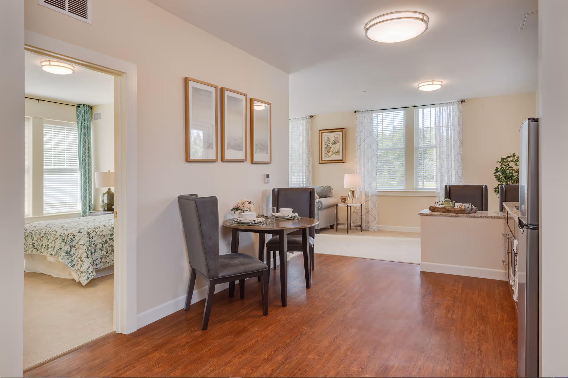 Interior view of a senior living apartment at The Residence at Penniman Hill showing a small dining area with a round table and two chairs, a living room with a sofa and side table with a lamp, large windows with sheer curtains, and a partial view of a bedroom with a bed covered in a floral bedspread. The flooring is a mix of wood and carpet, and the walls are decorated with framed artwork.