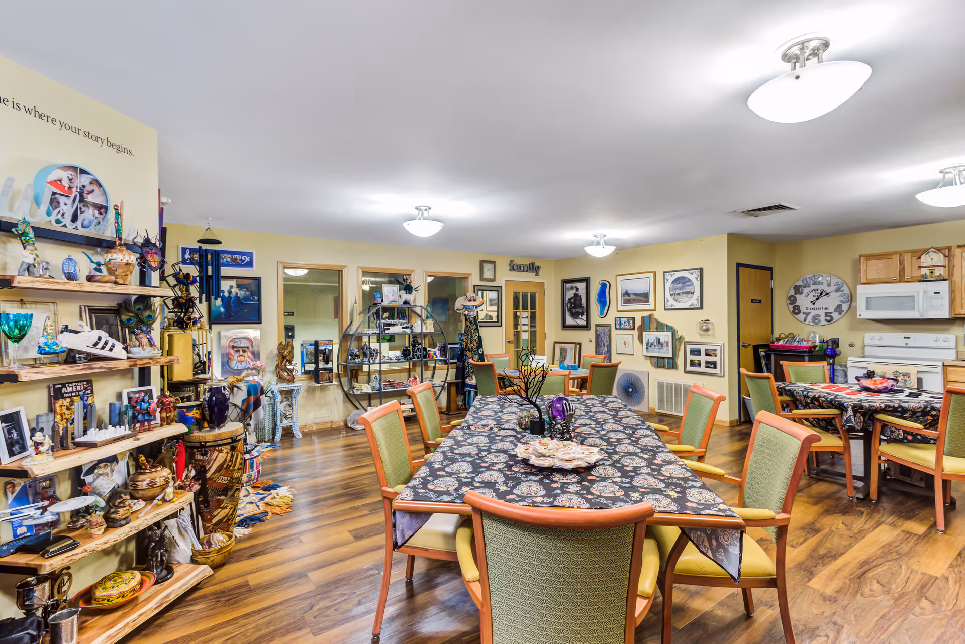 A cozy and colorful common area in a senior living facility featuring multiple tables covered with patterned tablecloths and surrounded by chairs. The room has wooden flooring and walls adorned with various framed pictures and decorative items. On the left side, there are shelves filled with collectibles and memorabilia. The back wall includes a kitchen area with a stove, microwave, and a large clock. The space is well-lit with ceiling lights.