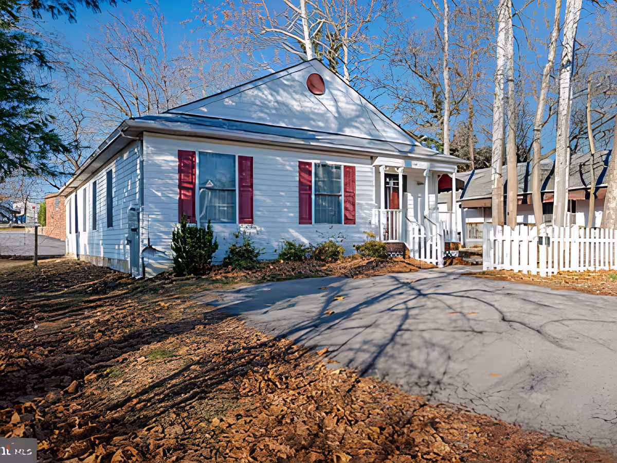 Exterior view of a single-story white house with red shutters and a small front porch with white railing. The house is surrounded by bare trees and a white picket fence, with a paved driveway in front and dry, brown soil on the sides.