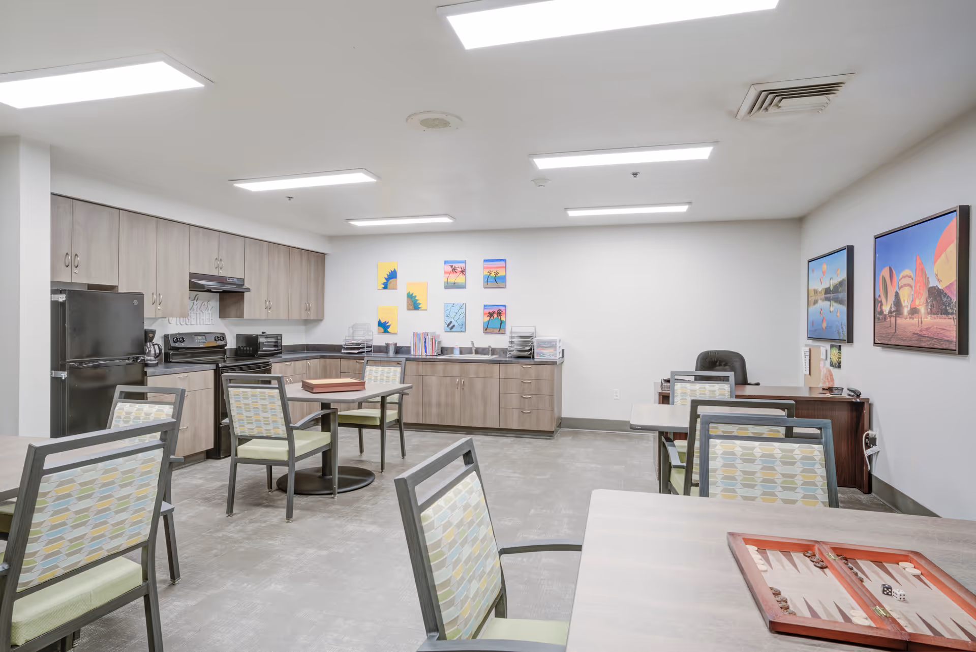 A bright, spacious room with a kitchenette featuring wooden cabinets, a black refrigerator, stove, and microwave. Several tables and chairs with patterned upholstery are arranged around the room. The walls are decorated with colorful artwork and framed pictures of hot air balloons. A desk with a black office chair is positioned against one wall.