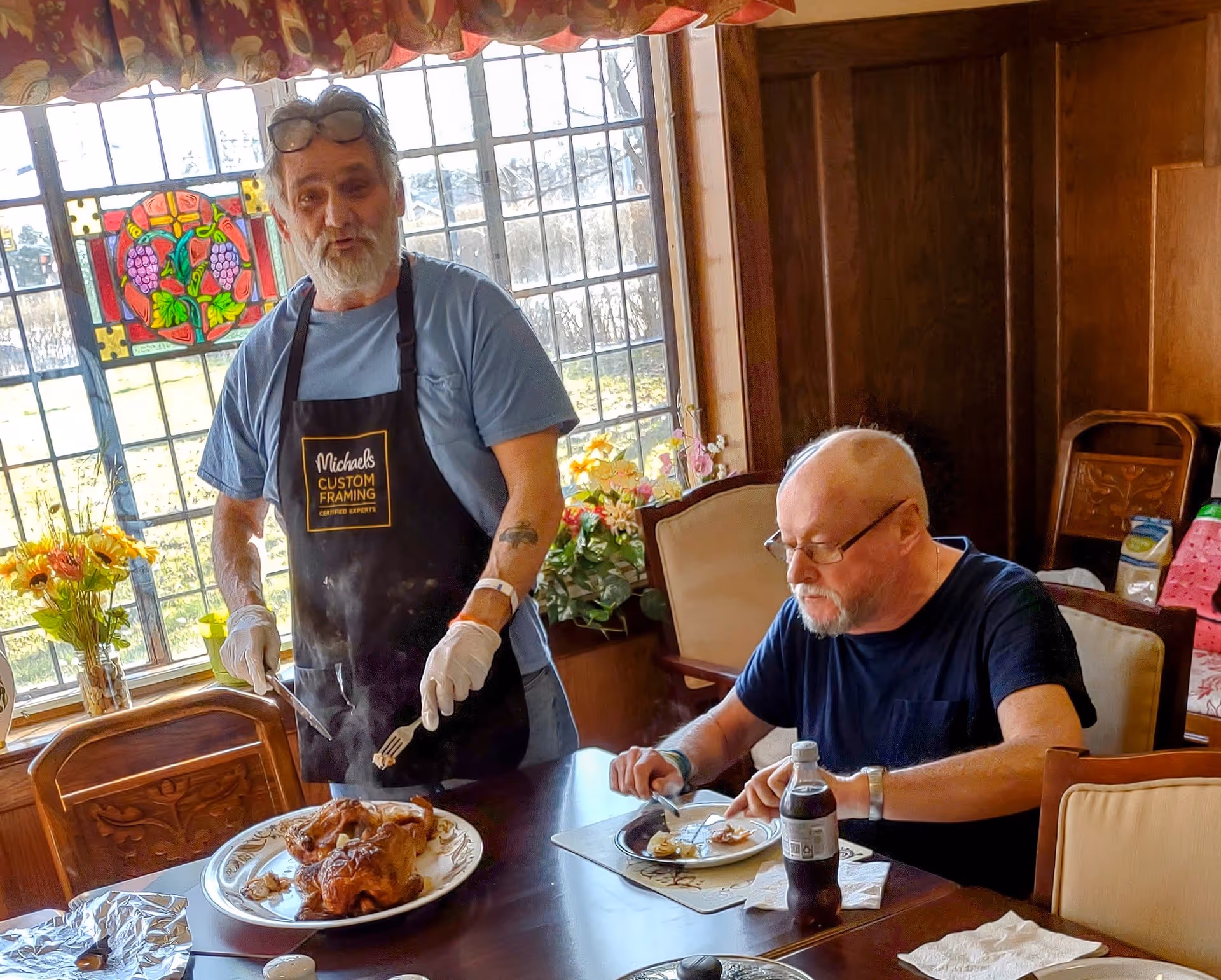 Two elderly men in a dining room with wooden paneling and a large window with stained glass. One man wearing a blue shirt and a black apron is carving a roasted chicken on a plate, while the other man with glasses and a black shirt is seated at the table eating. The table has placemats, a bottle of soda, and napkins.