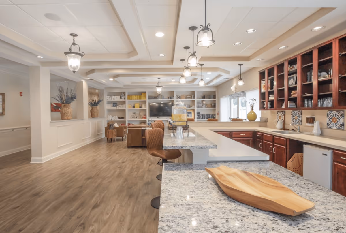 Open communal kitchen and lounge with granite countertops, bar seating, pendant lights, and a seating area with shelving in the background.