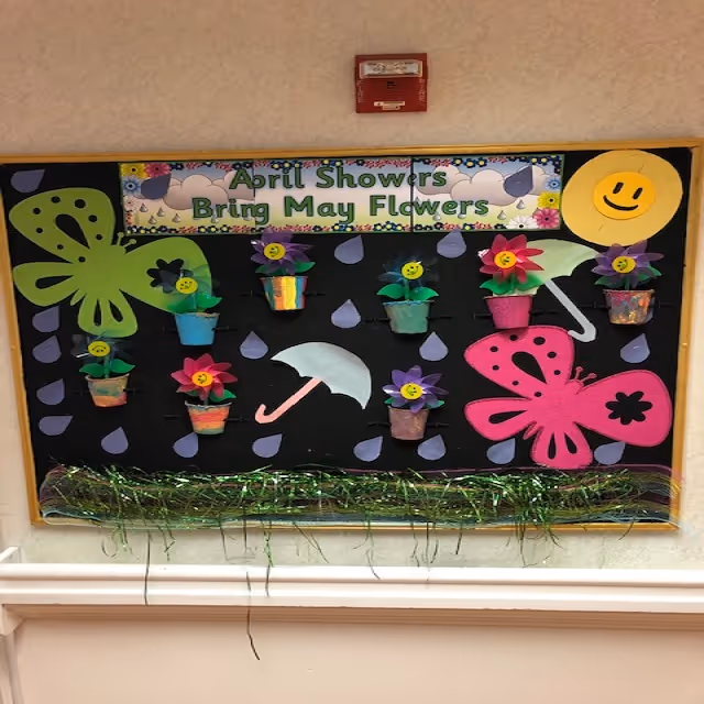 A colorful bulletin board decorated with paper flowers in small pots, two large butterflies, an umbrella, raindrops, and a smiling sun. The board has a sign that reads 'April Showers Bring May Flowers.' Green decorative grass is attached along the bottom edge of the board.