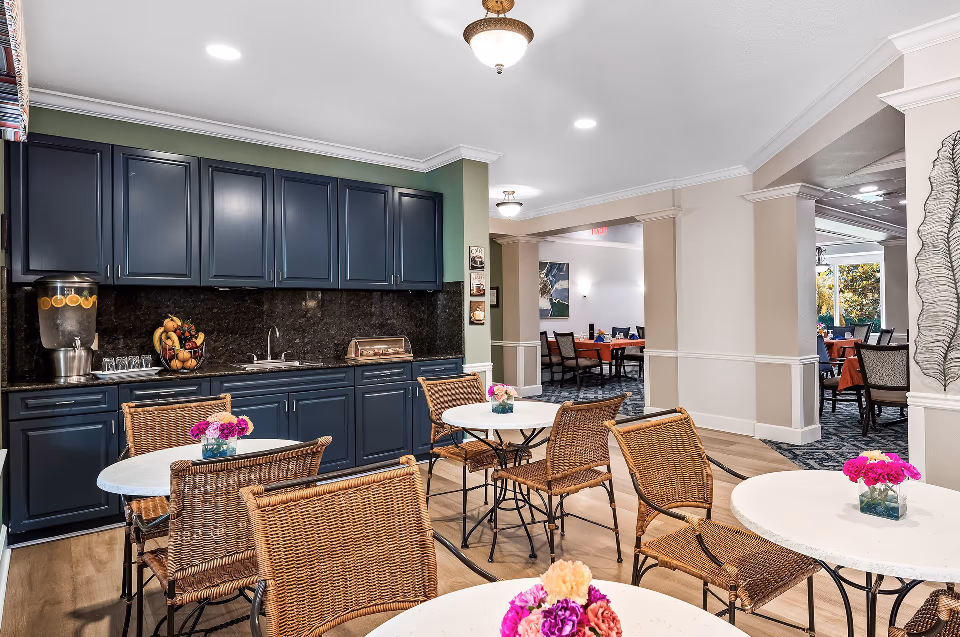 Interior view of a dining area in a senior living facility featuring round white tables with wicker chairs. Each table has a small vase with colorful flowers. In the background, there is a kitchenette with dark blue cabinets, a sink, a water dispenser with lemon slices, and a fruit basket. The room is well-lit with ceiling lights and has a neutral color scheme with green and beige walls. Additional dining tables and chairs are visible through an open doorway.