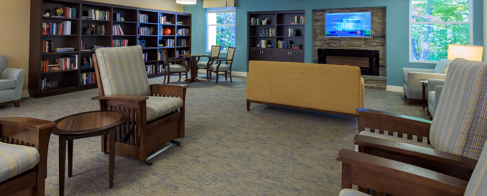 A cozy living room area with wooden armchairs upholstered in striped fabric, a small wooden side table, a beige sofa facing a wall-mounted TV above a stone fireplace, large windows letting in natural light, and dark wood bookshelves filled with books along the walls.