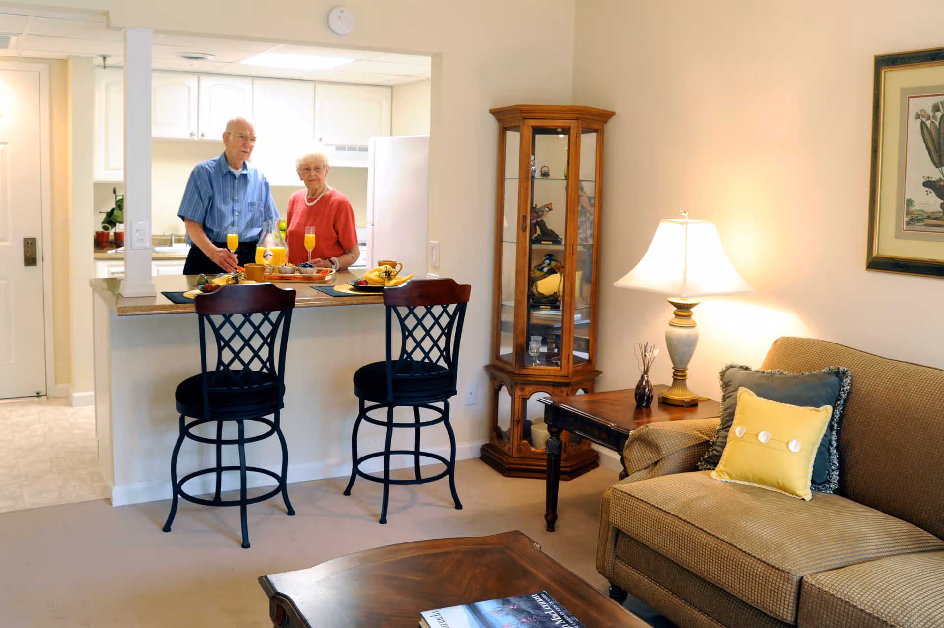 An elderly couple standing behind a kitchen counter in a retirement community apartment. The kitchen has white cabinets and appliances. In the foreground, there are two black bar stools at the counter, a wooden coffee table, a beige couch with decorative pillows, a side table with a lamp, and a wooden display cabinet with glass doors. The room is warmly lit and decorated with a framed picture on the wall.