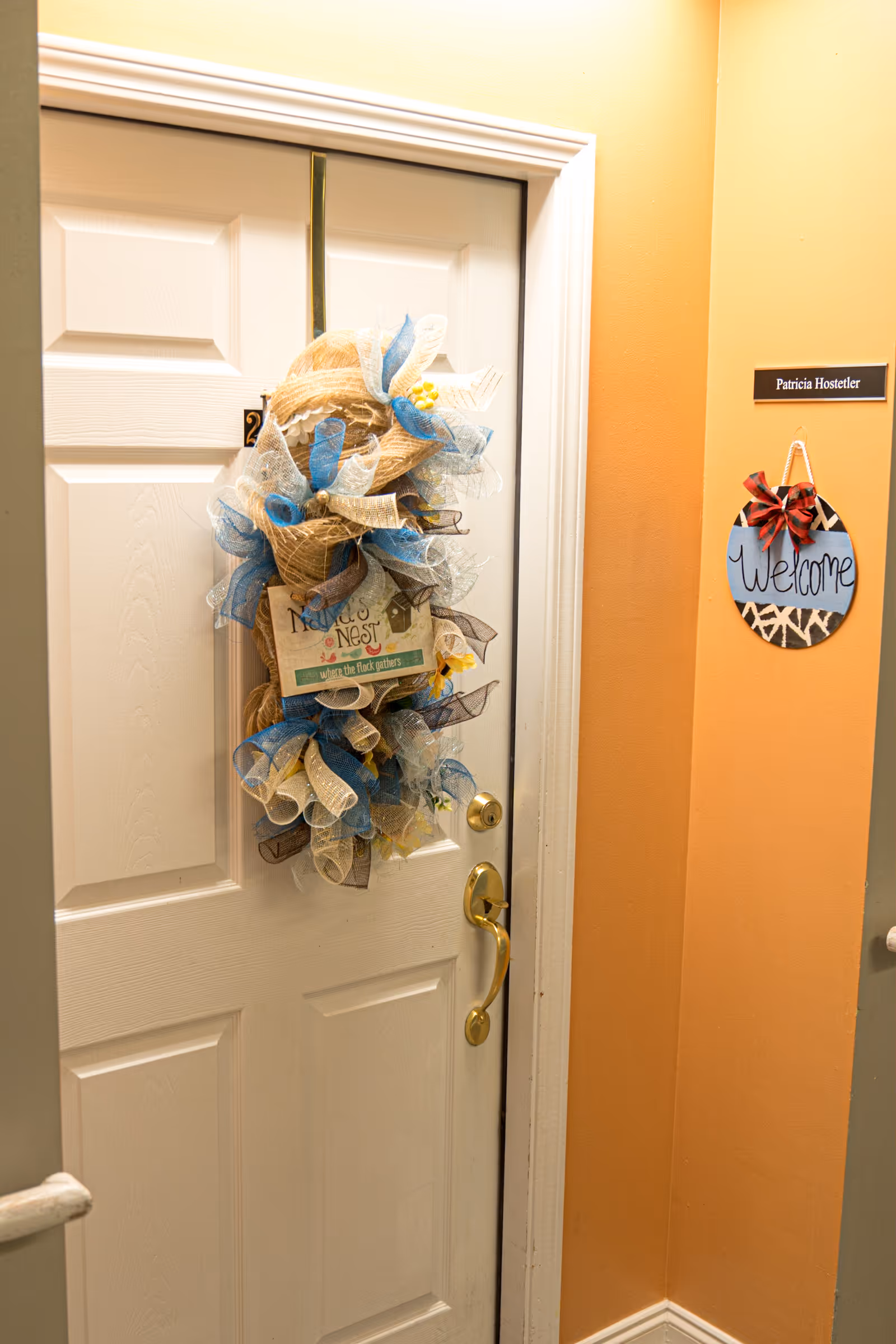 A white door decorated with a large decorative wreath made of beige, blue, and white mesh ribbons. The wreath has a small sign that reads 'Robin's Nest' and some small yellow flowers. To the right of the door, there is a wall with a nameplate that says 'Patricia Hostetler' and a round hanging sign with a red bow that says 'Welcome'. The walls are painted a warm orange color.