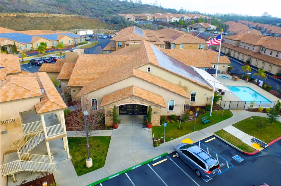 Aerial view of the Ponté Palmero senior living facility showing multiple beige buildings with orange tiled roofs, a parking lot with cars, a swimming pool enclosed by a fence, an American flag on a flagpole, and surrounding greenery and pathways.