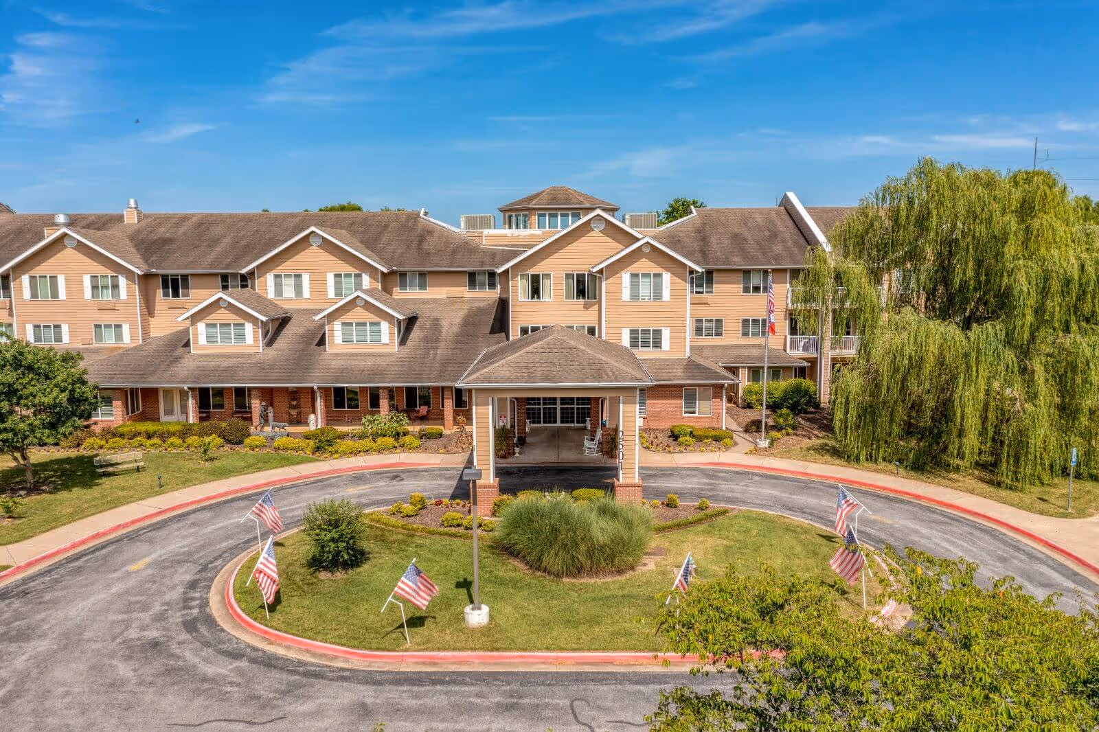 Front exterior view of Morada Rogers, a multi-story senior living facility with beige siding and a covered entrance. The building is surrounded by a circular driveway with American flags displayed on the grassy island in the center. There are trees and shrubs around the building under a clear blue sky.