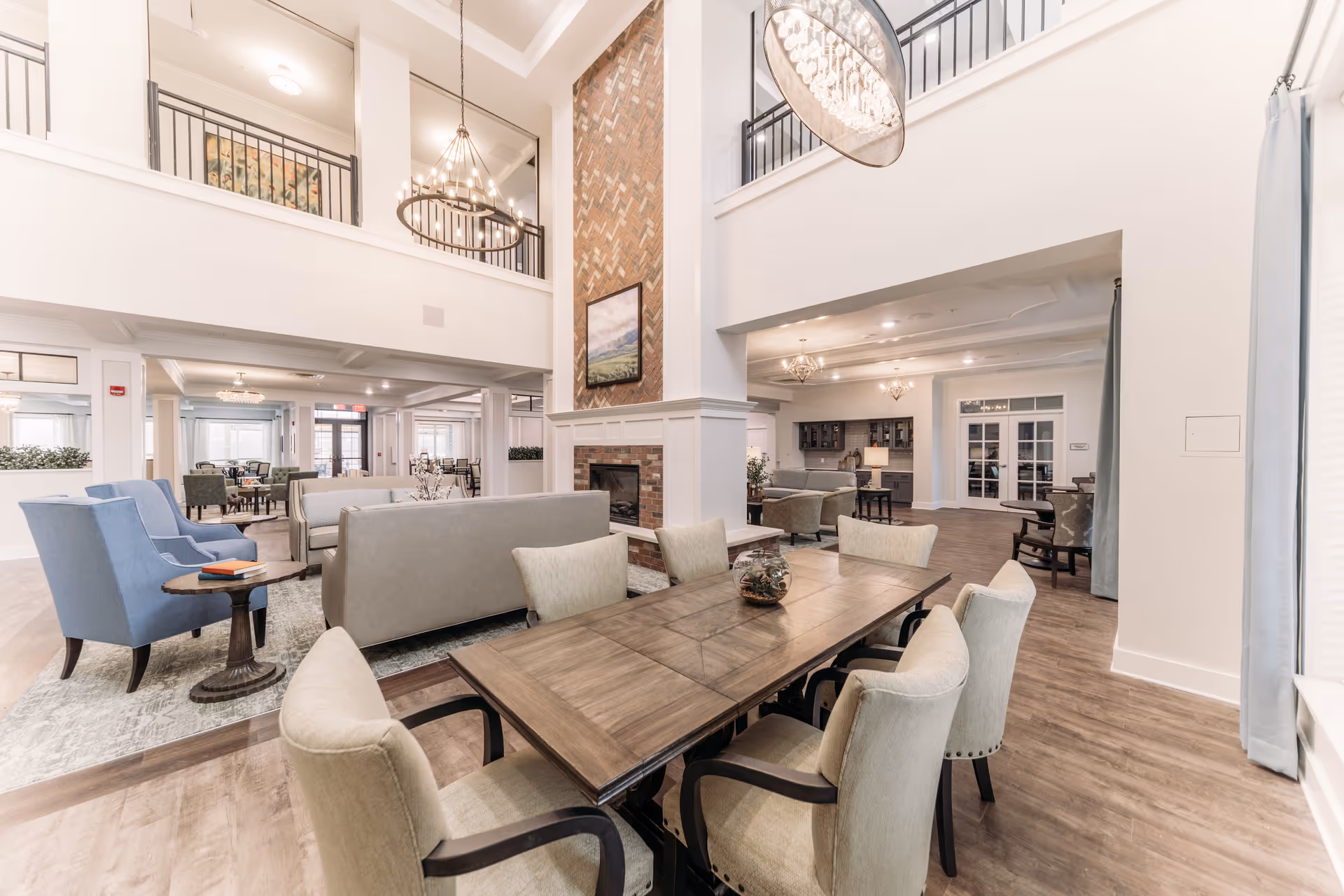 Spacious and well-lit senior living facility common area with a large wooden dining table surrounded by beige upholstered chairs in the foreground. Behind the table, there are multiple seating areas with sofas and armchairs arranged around a central brick fireplace with a painting above it. The room features high ceilings with modern chandeliers, light-colored walls, wood flooring, and large windows with curtains allowing natural light to fill the space.