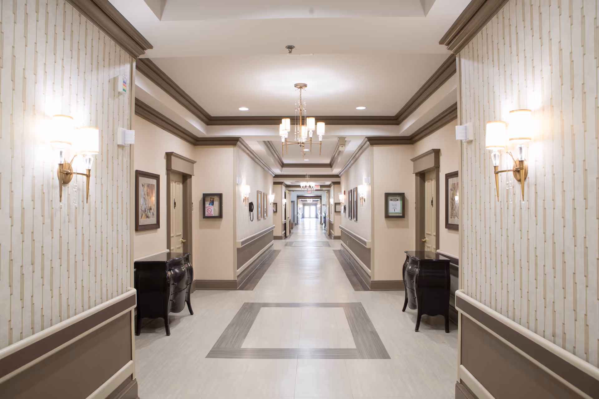 A long, well-lit hallway in a senior living facility with beige walls, decorative wall sconces, framed artwork, and small black cabinets along the sides. The ceiling has recessed lighting and chandeliers, and the floor features a patterned design.