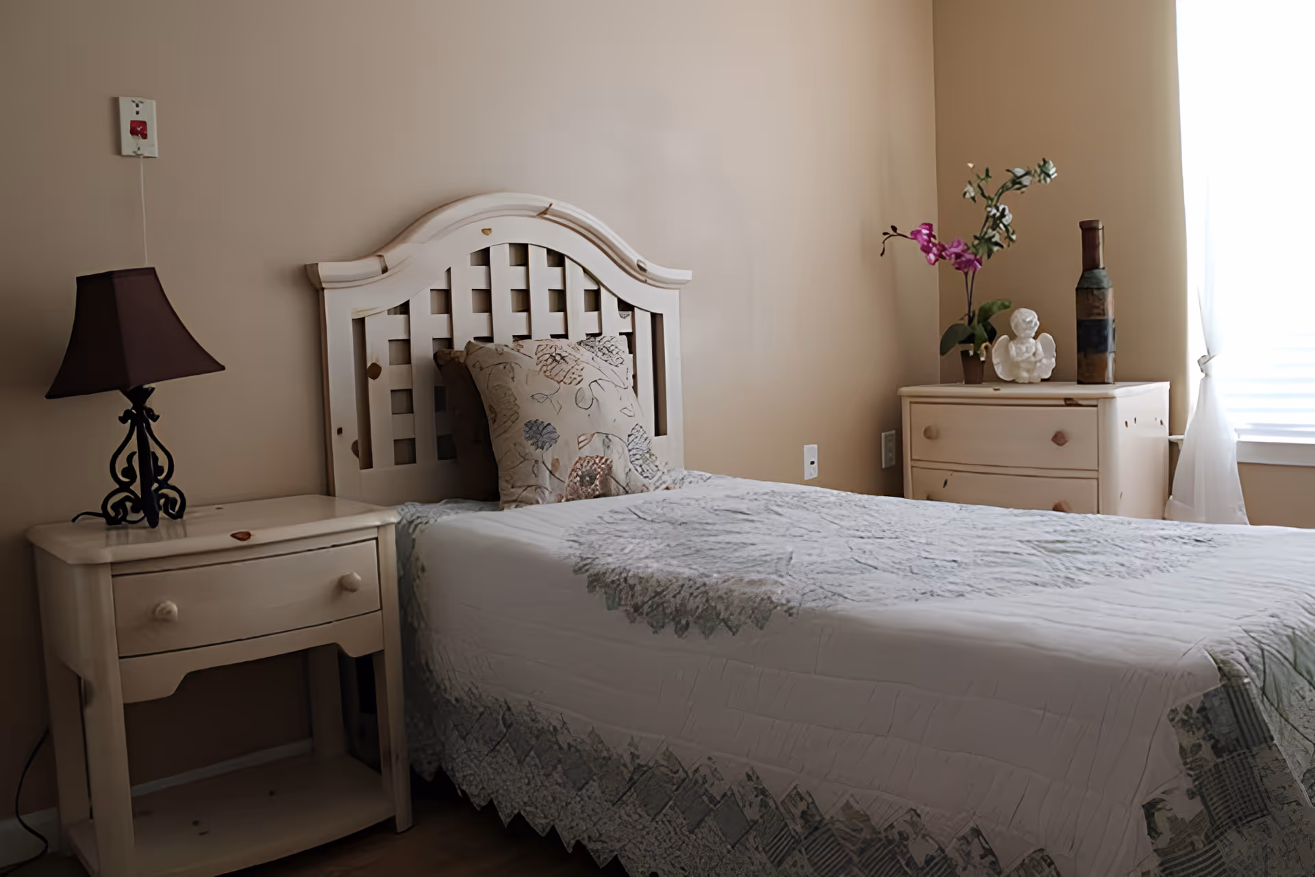 A cozy bedroom with a single bed featuring a white wooden headboard and a quilted bedspread. There are two pillows on the bed. On the left side of the bed is a small wooden nightstand with a dark brown lamp. On the right side, there is a wooden dresser with decorative items including a potted orchid, a small angel figurine, and a tall bottle. A window with sheer white curtains allows natural light to brighten the room.
