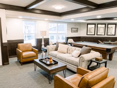 A well-lit common area featuring a beige tufted sofa with multiple cushions, two brown leather armchairs, a wooden coffee table with decorative items, and a pool table in the background. The room has large windows, dark wood paneling on the walls, framed artwork, and recessed ceiling lights.