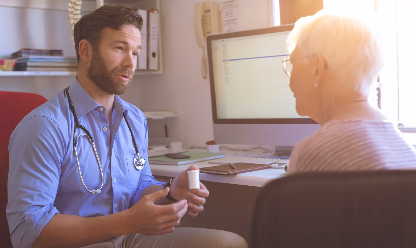 A male doctor with a stethoscope around his neck is sitting and talking to an elderly woman in an office setting. The doctor is holding a prescription bottle and explaining something to the woman, who is seated across from him. A computer monitor and office supplies are visible in the background.