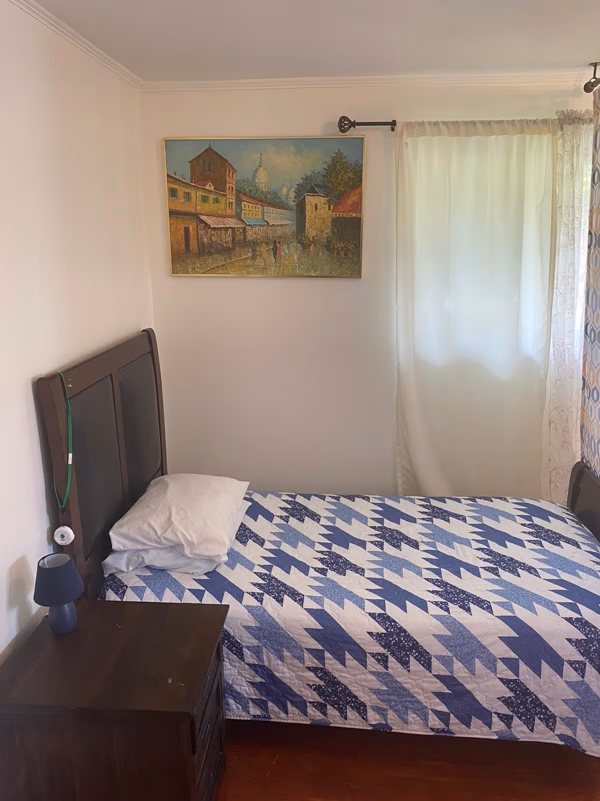 A small bedroom with a single bed covered in a blue and white patterned quilt. There is a white pillow on the bed. Next to the bed is a dark wooden nightstand with a small blue lamp on it. A painting depicting a street scene with buildings and people hangs on the wall above the bed. A window with white curtains is on the right side of the room.