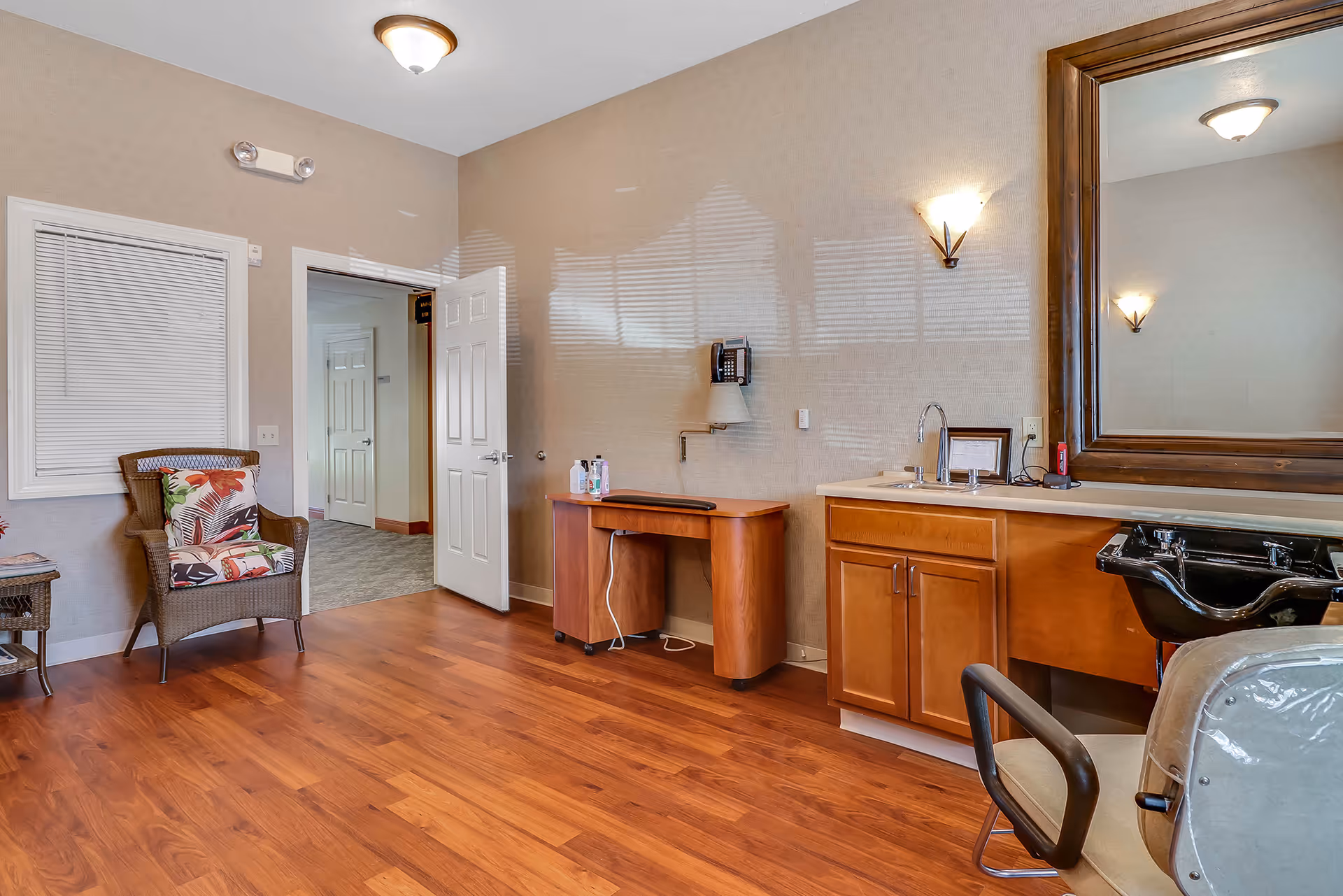 Interior room with wooden flooring, a wicker chair with floral cushions, a small wooden table, a wooden cabinet with a sink, a large mirror above the sink, a black salon-style wash basin, and a white door leading to a hallway.