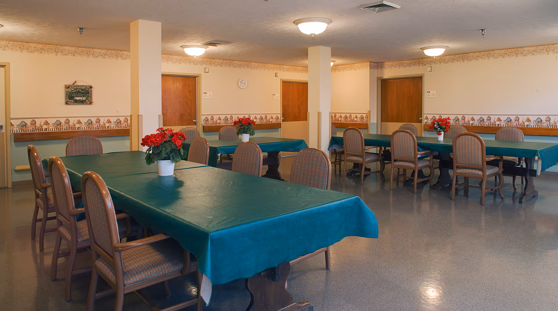 A dining room with several rectangular tables covered with green tablecloths, each table adorned with a small potted plant with red flowers. The room has beige walls with a decorative border featuring a village scene, wooden doors, and ceiling lights providing warm illumination.