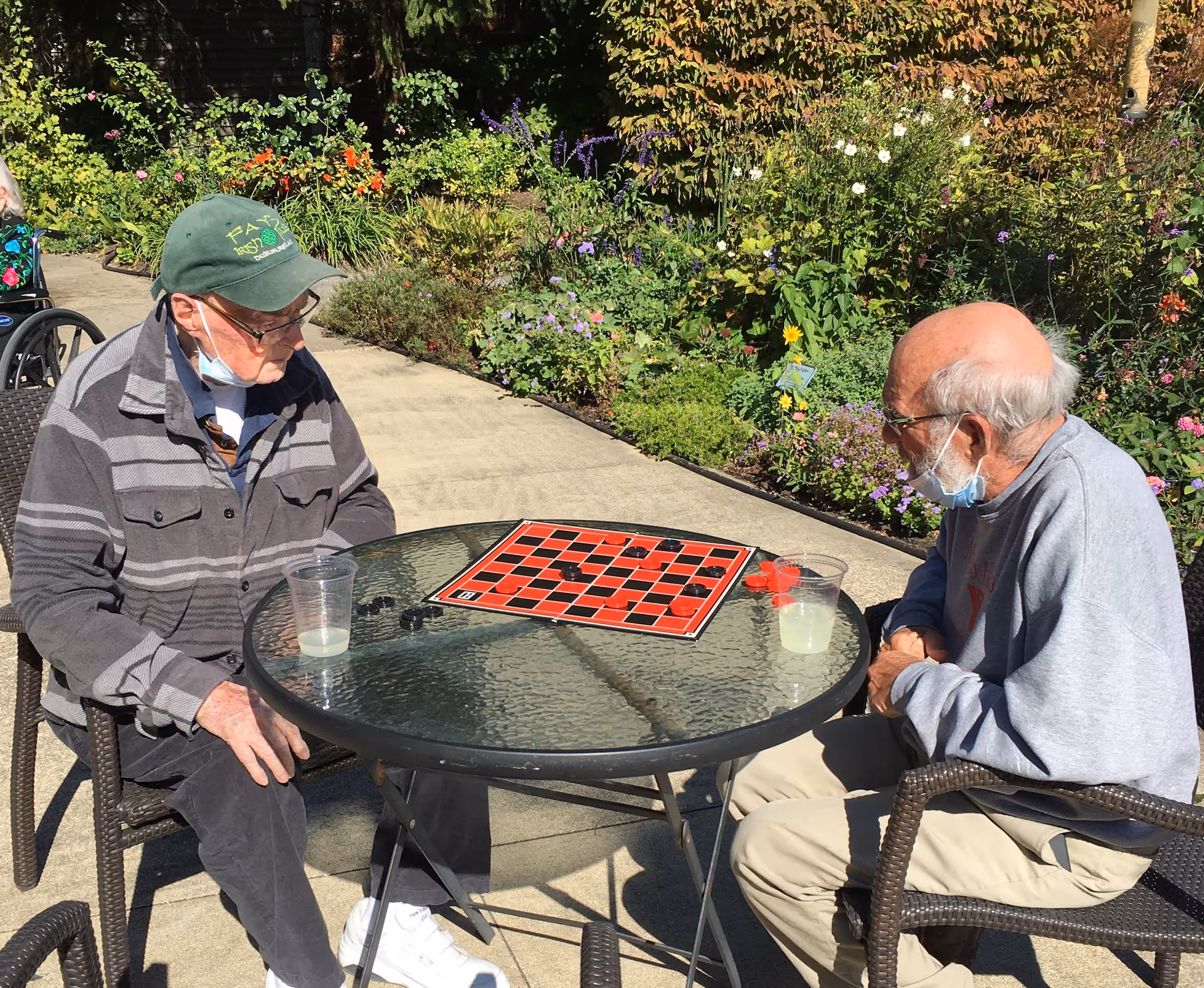 Two elderly men wearing masks sit at a round patio table playing checkers beside a flower garden.