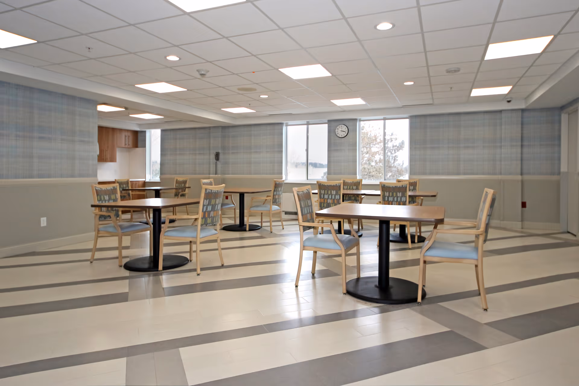 A bright dining room with several wooden tables and chairs arranged neatly. The room has large windows letting in natural light, a clock on the wall, and a patterned floor with gray and white stripes. The walls are decorated with light blue and gray wallpaper, and the ceiling has recessed lighting panels.