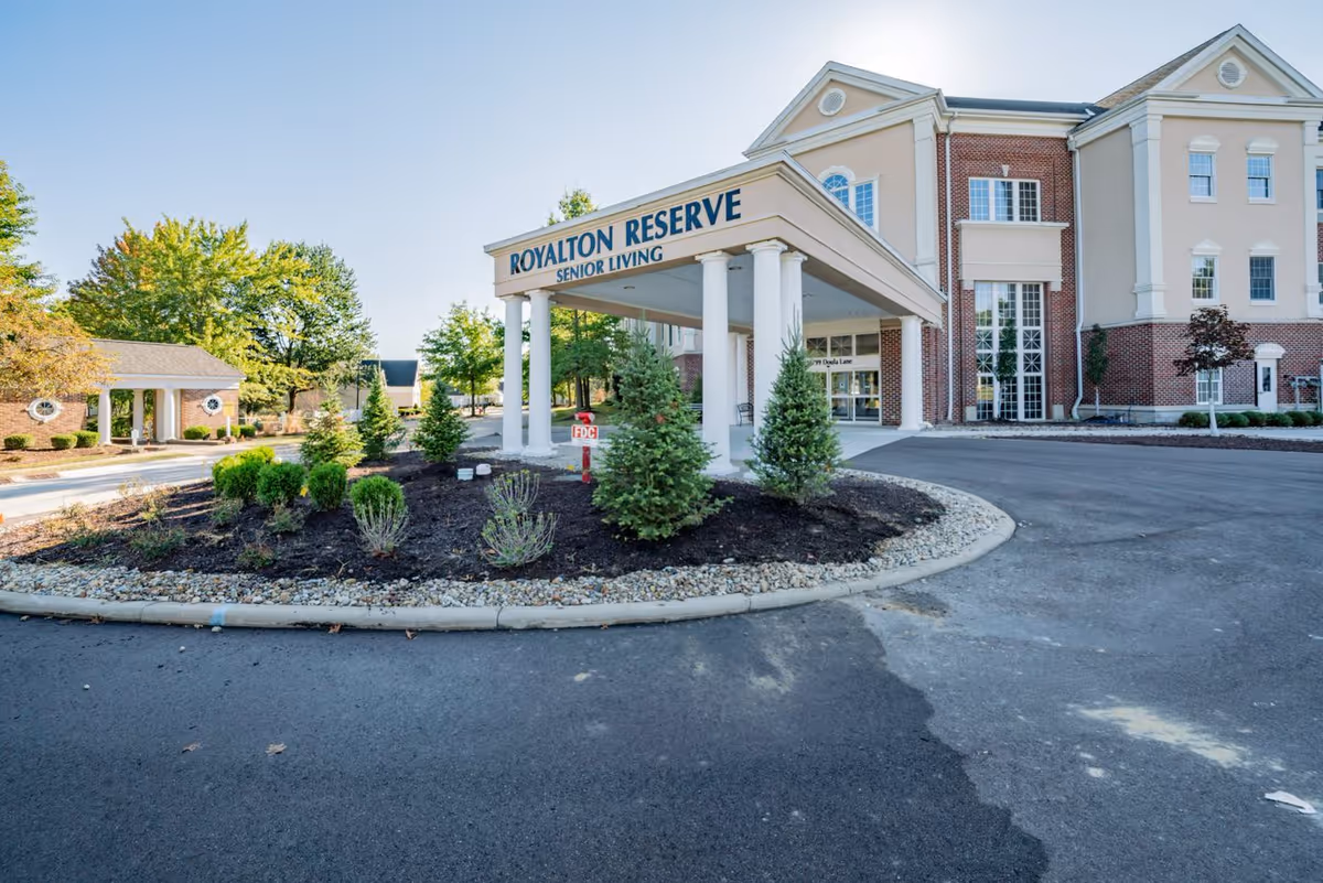 Exterior view of the Royalton Reserve Senior Living facility entrance with a covered drop-off area supported by white columns, landscaped garden beds with small trees and shrubs, and a clear blue sky.