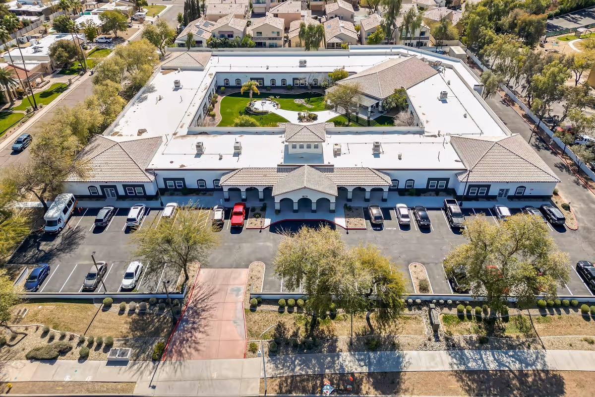 Aerial view of Inspirations Of Tempe senior living facility showing a large, single-story building with a central courtyard featuring green grass, trees, and walking paths. The building has a white roof and beige walls with a parking lot in front containing several cars. Surrounding the facility are residential houses and streets.