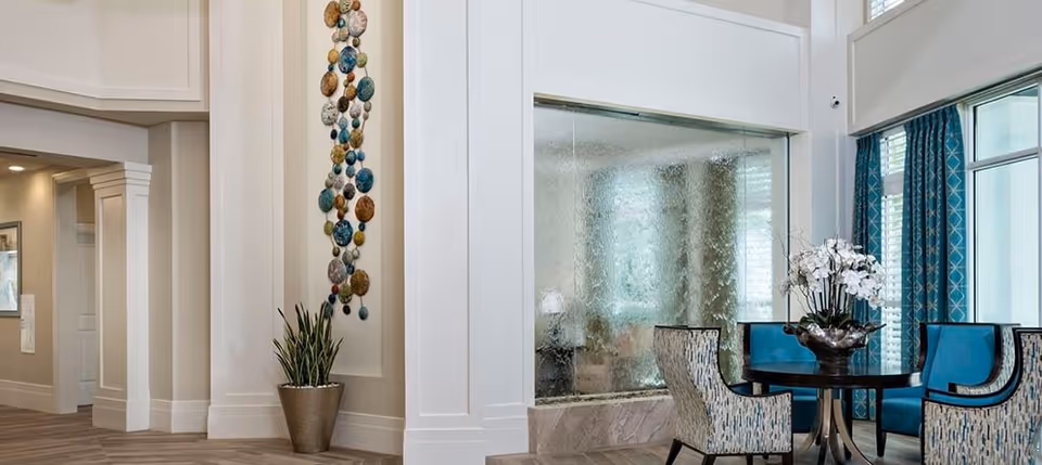 A bright and elegant interior common area with a round table surrounded by four upholstered chairs in blue and patterned fabric. Behind the table is a large textured glass panel with a water feature. To the left, there is a tall potted plant and a decorative wall hanging made of colorful circular elements. Large windows with blue patterned curtains allow natural light to fill the space.