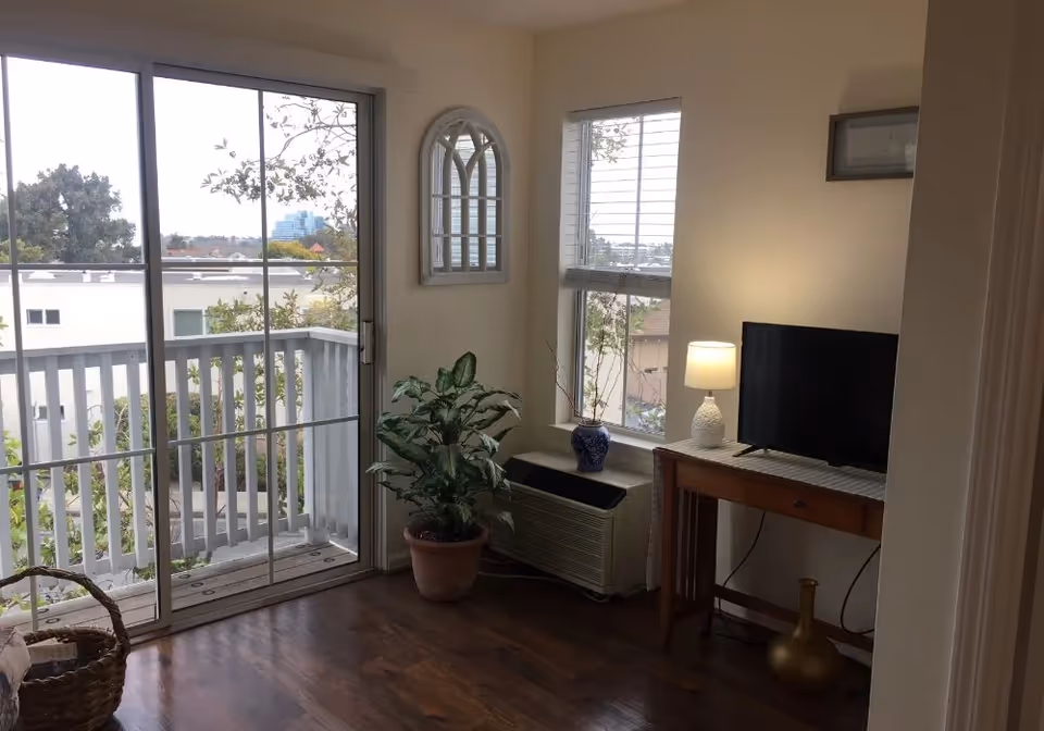 Cozy living area with sliding glass doors to a balcony, a potted plant, window unit, TV on a wooden console and a lit table lamp.