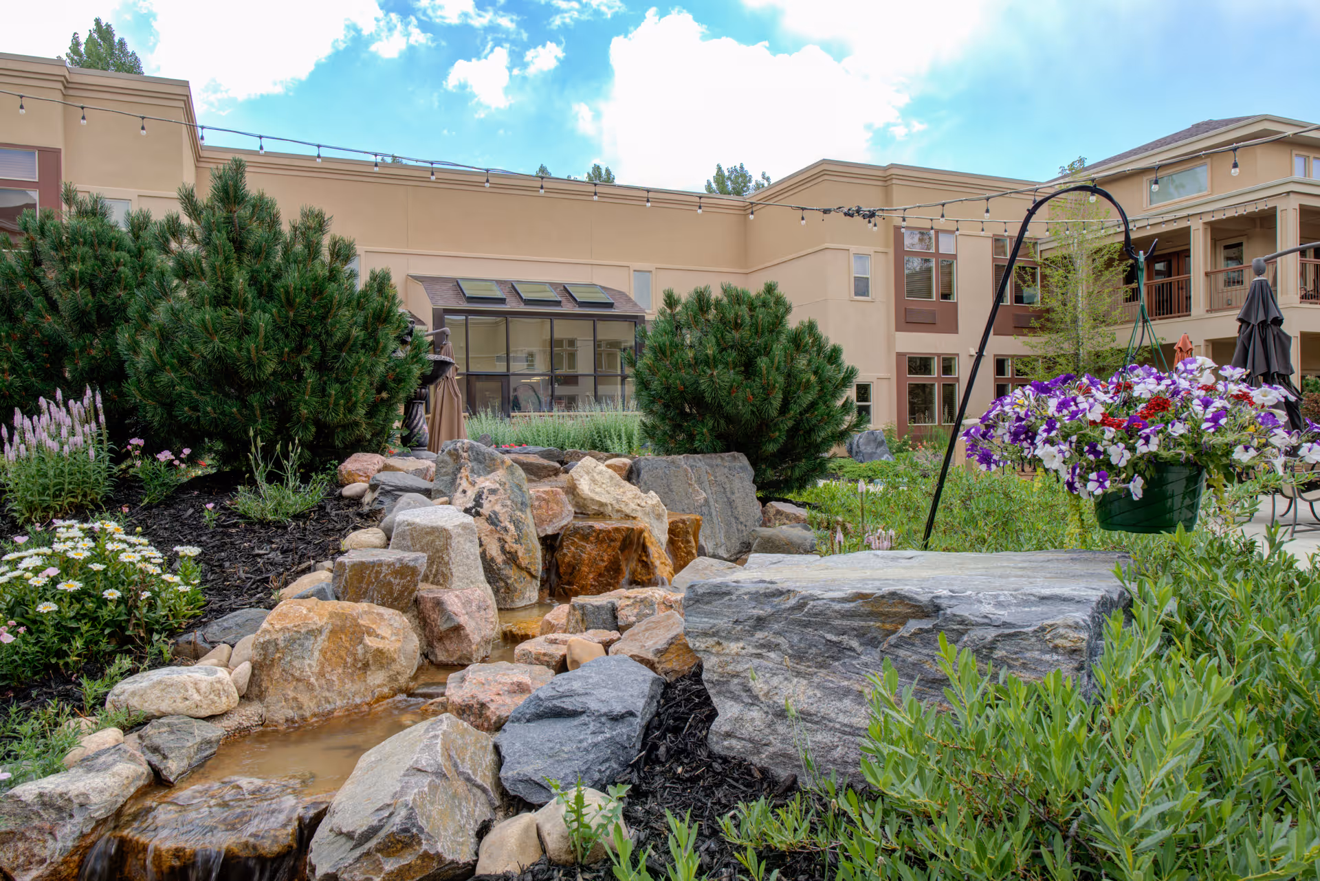 A landscaped outdoor garden area at the Gardens on Quail facility featuring a small rock water feature surrounded by various plants and flowers, with a hanging basket of purple and white flowers in the foreground. The beige building with large windows and string lights is visible in the background under a partly cloudy sky.