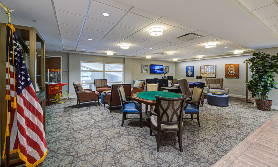 Spacious communal lounge with a card table, multiple seating areas, a TV, an American flag, and a popcorn cart.