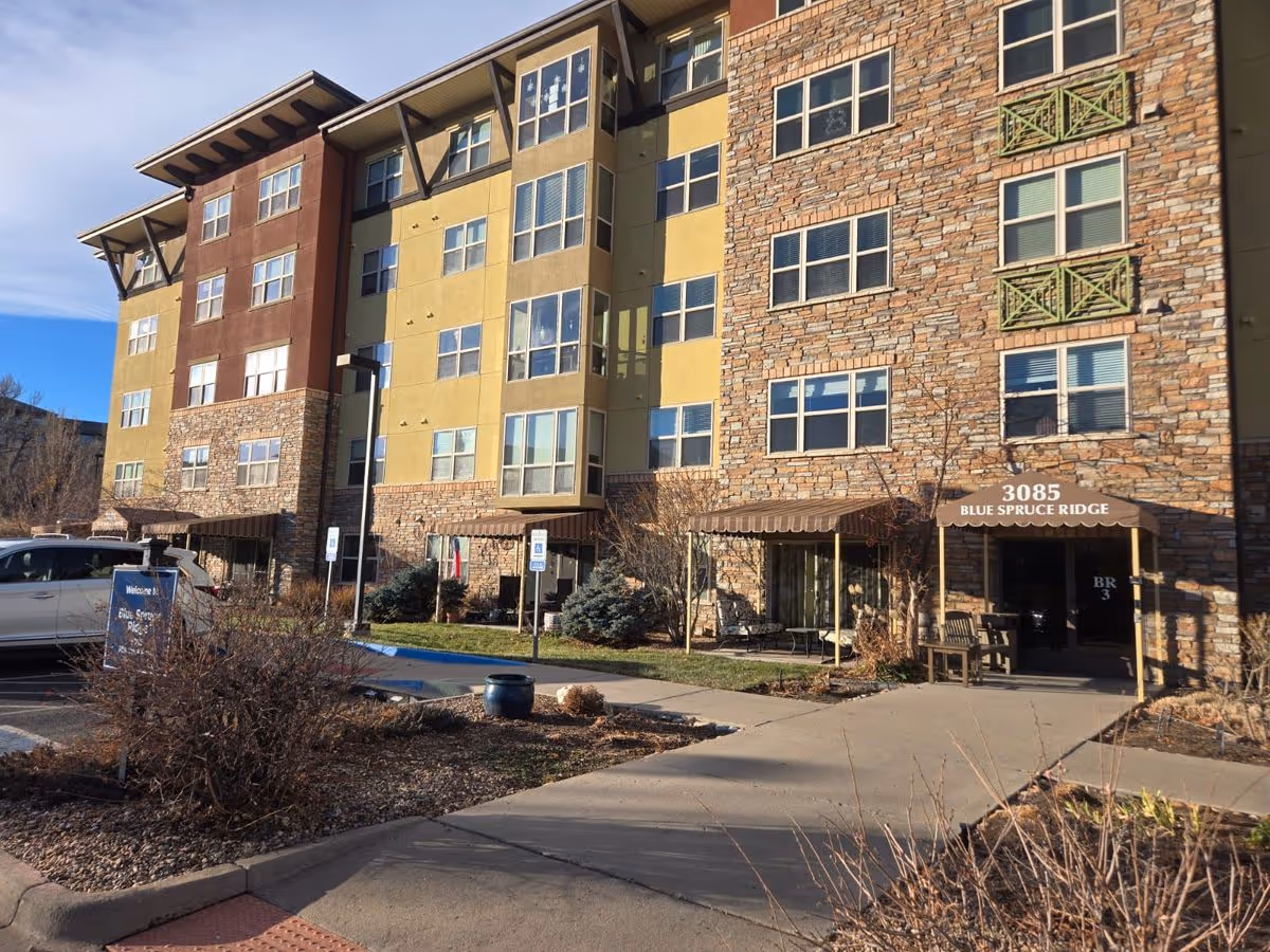 Exterior front of a multi-story senior living building with a stone facade, entrance canopy labeled "3085 Blue Spruce Ridge", landscaping and a parking area.