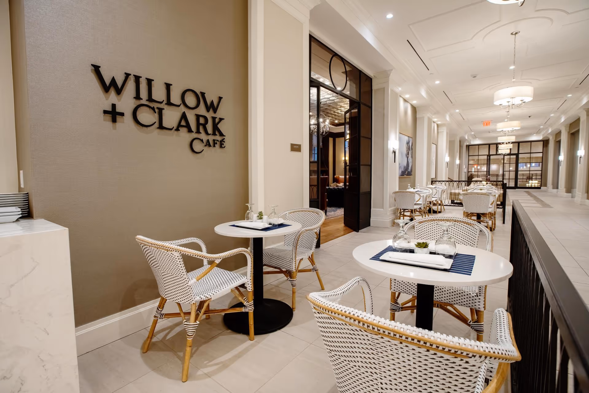 Bright indoor cafe with white wicker chairs and small round tables beside a wall sign reading "WILLOW + CLARK CAFE".