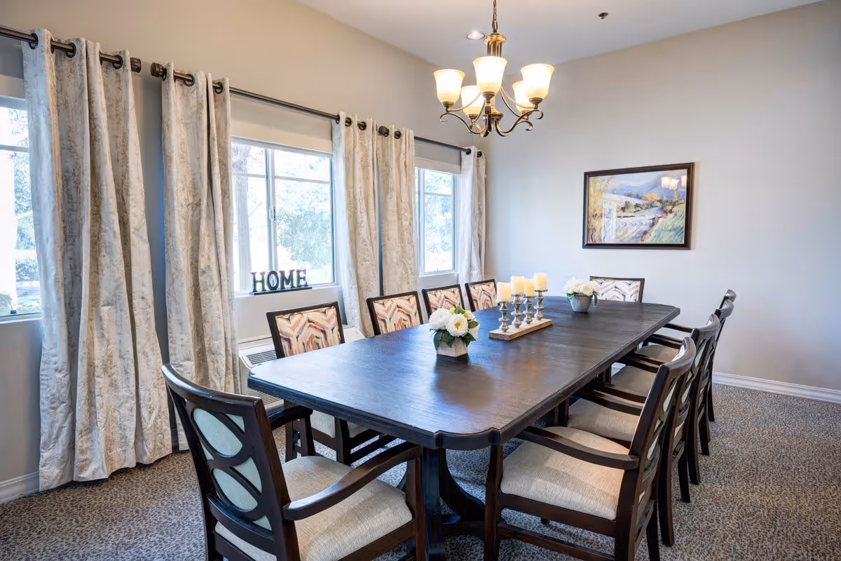 A dining room with a long dark wooden table surrounded by ten cushioned chairs. The table is decorated with two small flower arrangements and a candle holder with five candles. There are three large windows with light-colored curtains, and a decorative 'HOME' sign is placed on the windowsill. A chandelier with five lights hangs above the table, and a framed painting is on the wall.