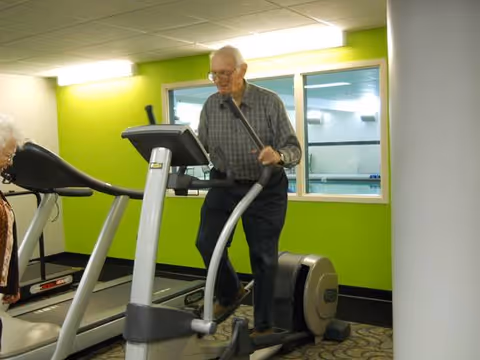 An elderly man exercising on an elliptical machine in a fitness room with bright green walls and a window showing an adjacent room. Another elderly person is partially visible near a treadmill.
